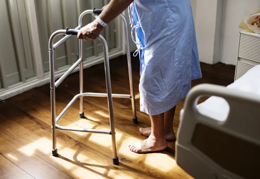 Residente caminhando com andador e apoio ao lado da cama hospitalar